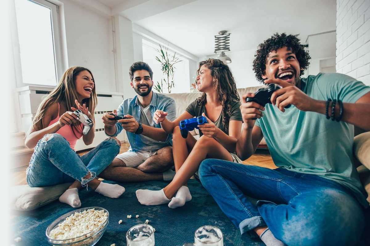 Four Friends Sitting On The Floor Playing Video Games, Laughing. Popcorn And Beverages Are Visible in The Foreground